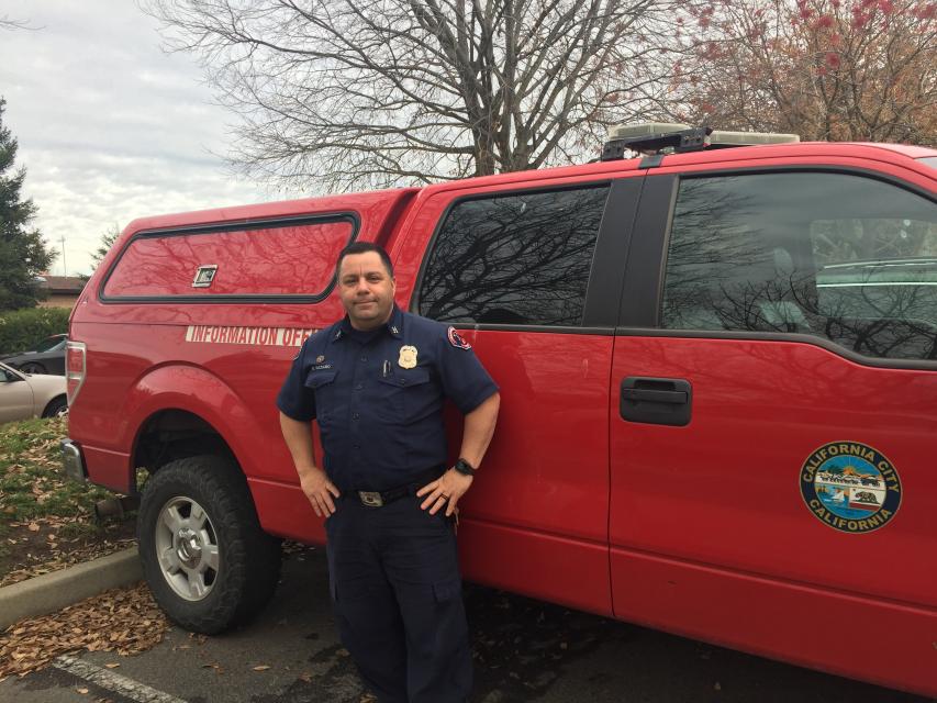 Officer, in uniform, posing in front of a red truck on a cloudy, fall day.