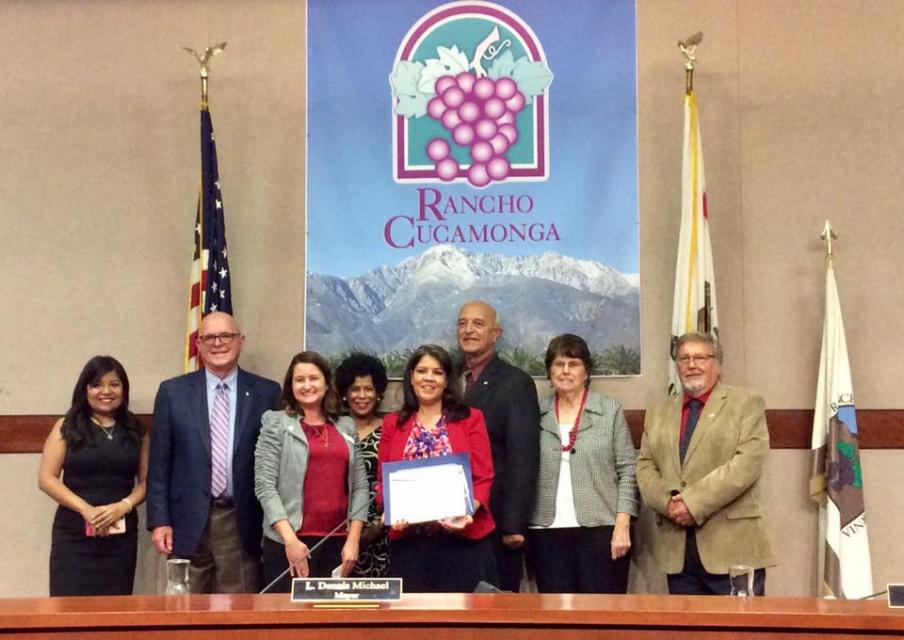 Francie Palmer holding award, surrounded by a group of people in a board room with a Rancho Cucamonga backdrop and flags.
